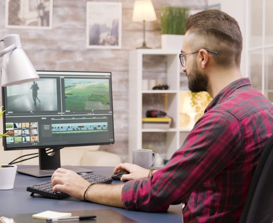 Creative filmmaker working on post production of a movie while working from home. Girlfriend in the background is walking in the house and talking on the phone.